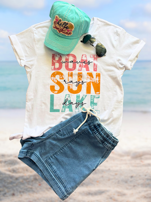 Boat Waves, Sun Rays, Lake Days V-neck tee with colorful text, turquoise Hello Sunshine cap, and denim shorts on a beach.