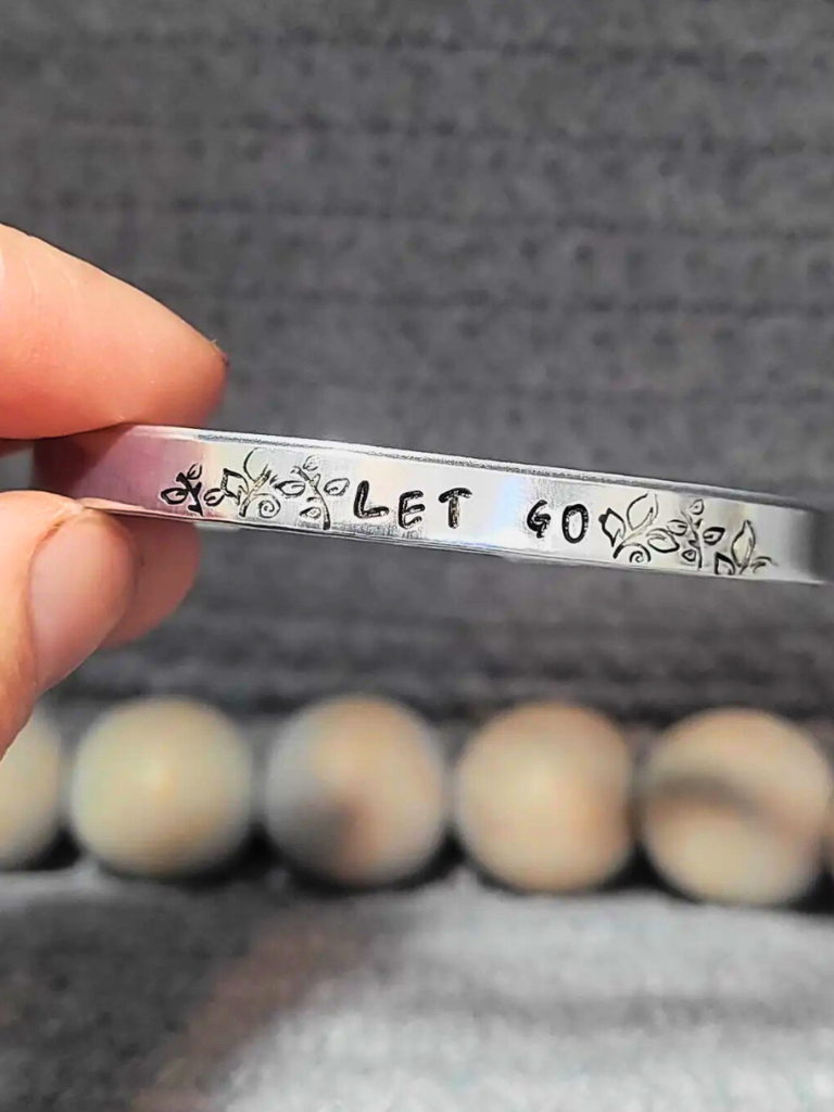 Let Go, aluminum bracelet with hand stamped text , sitting on a textured surface with wooden beads.