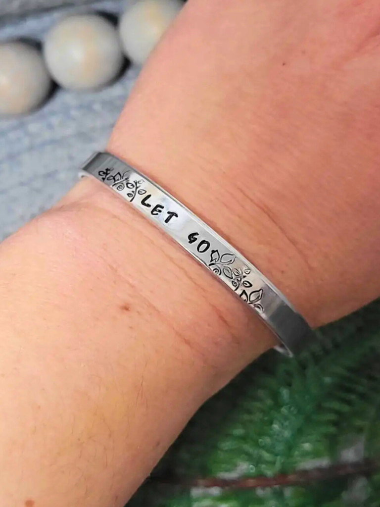 Let Go, aluminum bracelet with hand stamped text, on a person's wrist, on a textured surface with wooden beads and greenery.