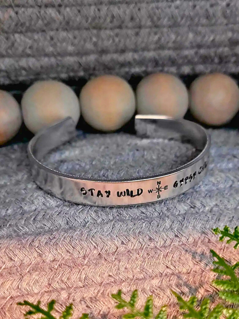 Stay Wild Gypsy Child aluminum bracelet with hand stamped text, sitting on a textured surface with wooden beads and greenery.