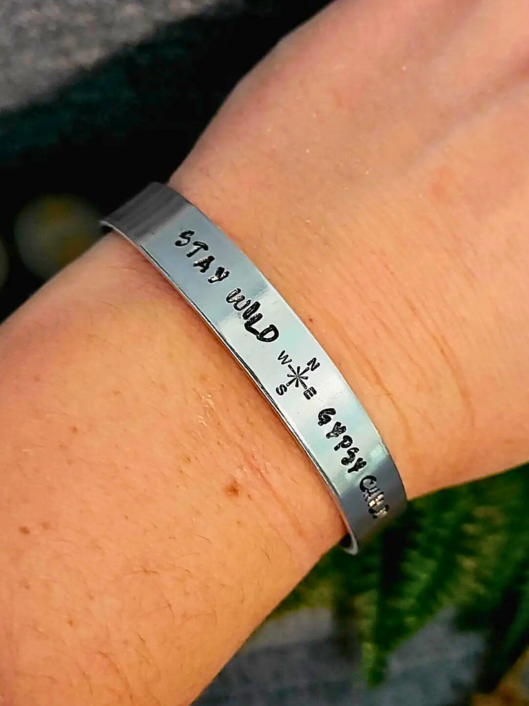 Stay Wild Gypsy Child aluminum bracelet with hand stamped text, sitting on a person's wrist with greenery in background.