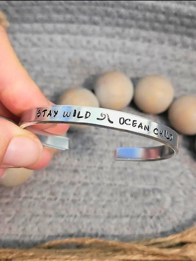 Stay Wild Ocean Child aluminum bracelet with hand stamped text, sitting on a textured surface with wooden beads in background.