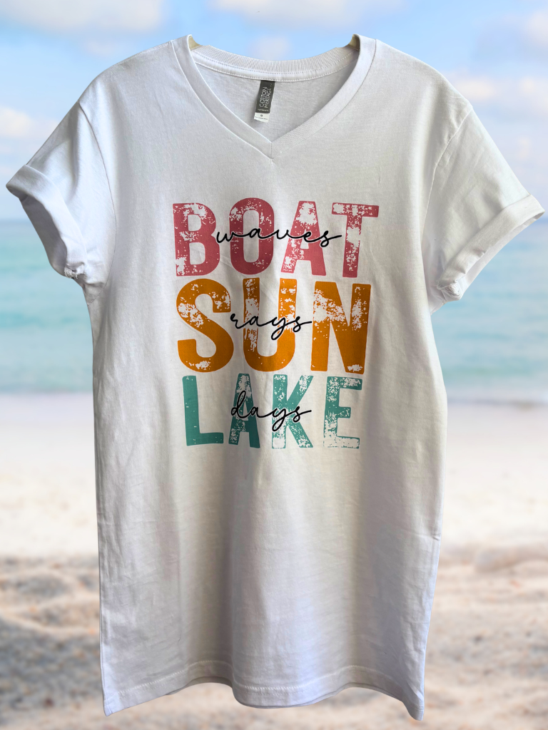 White V-neck Boat Wacces Sun Rays Lake Days t-shirt with colorful text design on a beach background Flat Lay Image