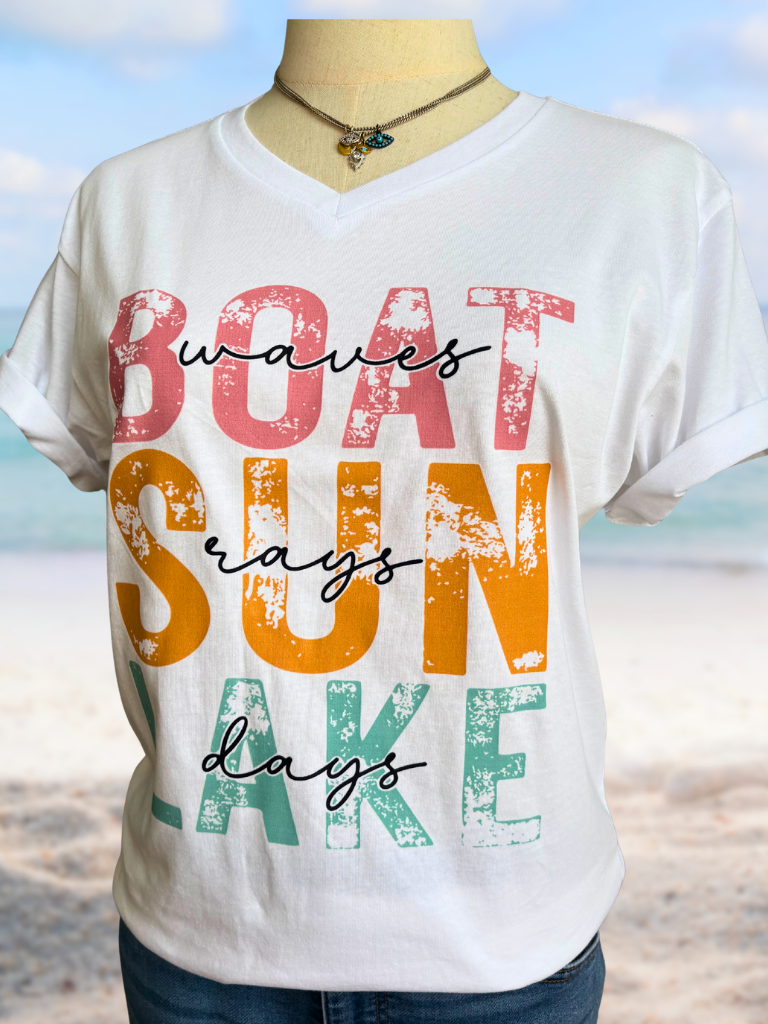 White v-neck t-shirt with colorful text 'Waves, Boat, Sun, Days' on a beach tucked into jeans, closer up with Eyes of Protection necklace