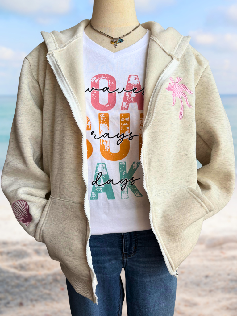 Mannequin wearing Coastal Bloom HIbiscus hoodie, white v-neck Boat Waves, Sun Rays, Lake Days tee with colorful text, and jeans against a beach background.