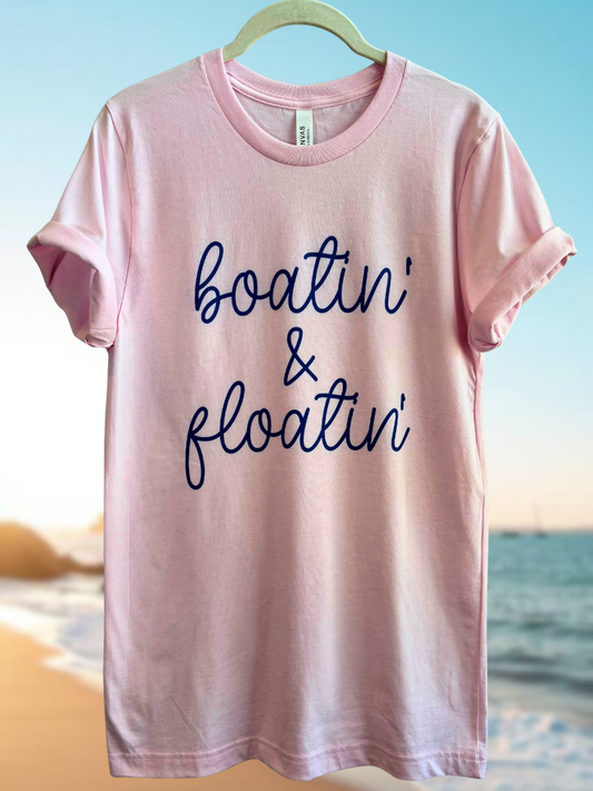 Pink t-shirt with 'Boatin' & Floatin' blue text on a hanger against a beach background.