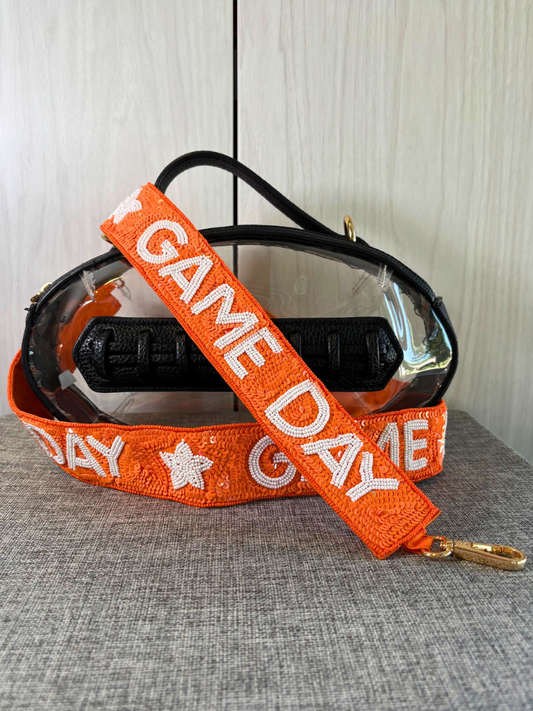 An orange woven strap with white 'GAME DAY' lettering and a gold clasp, draped over a clear plastic and black pouch against a light wood background.