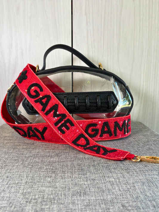 A clear plastic face shield with a black strap and a red glittery shoulder strap featuring black 'GAME DAY' text, displayed on a gray fabric surface against a light wood background.