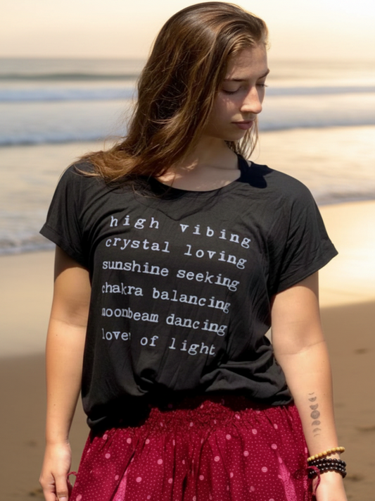 A woman wearing a black graphic t-shirt with white text that reads 'high vibing crystal loving sunshine seeking chaira balancing moonbeam dancing love of light'