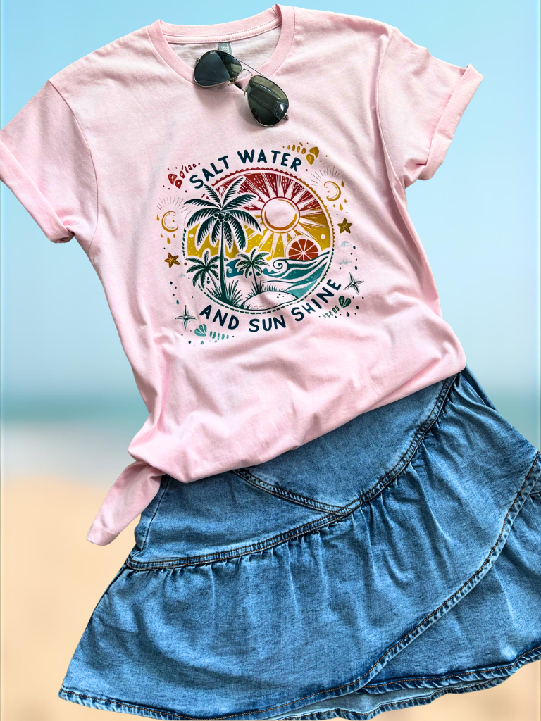 Salt Water and Sun Shine Tee, tropical design and sunglasses worn with our asymmetrical ruffle overlay, denim skirt by a beach.