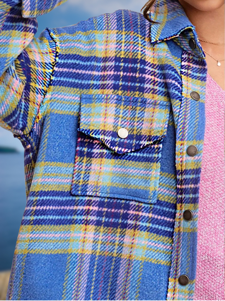 A close-up of a blue plaid button-up shirt worn over a pink knit top, featuring a chest pocket with a button flap and dark metal buttons.
