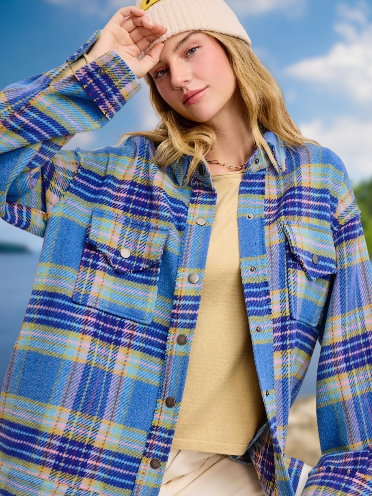 A woman wearing a blue, yellow, and purple plaid button-up flannel shirt over a beige ribbed knit top and a cream beanie, photographed outdoors against a blue sky background.