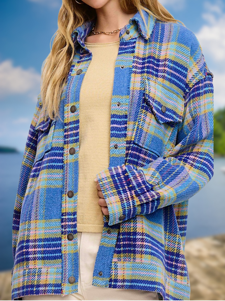 A woman wearing an oversized blue plaid button-up shirt over a beige top, photographed outdoors with a blurred sky and water background.