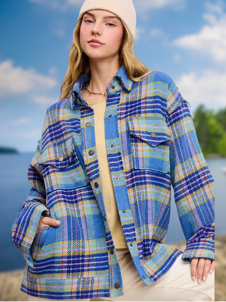 A woman wearing a blue, yellow, and  plaid pink button-up flannel shirt jacket over a beige top and pants, with a cream beanie, set against a scenic outdoor background of water and sky.