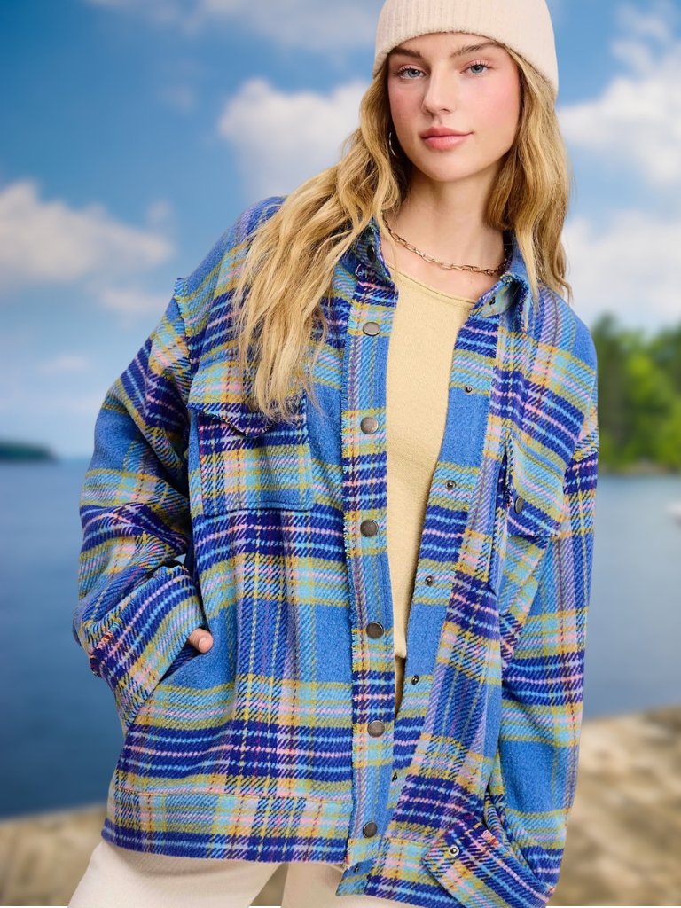 A woman wearing an oversized blue plaid button-up shirt jacket over a beige top, with a cream beanie, standing outdoors near water and trees.