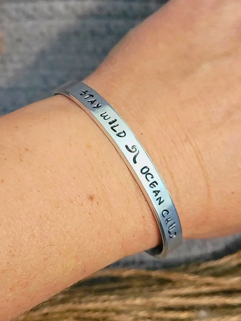 Stay Wild Ocean Child aluminum  bracelet with hand stamped text, sitting on a textured surface.