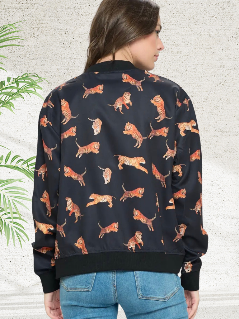 A woman viewed from the back wearing a black bomber jacket covered in an orange and beige tiger print, paired with blue jeans against a textured light gray wall with a green plant leaf on the left.