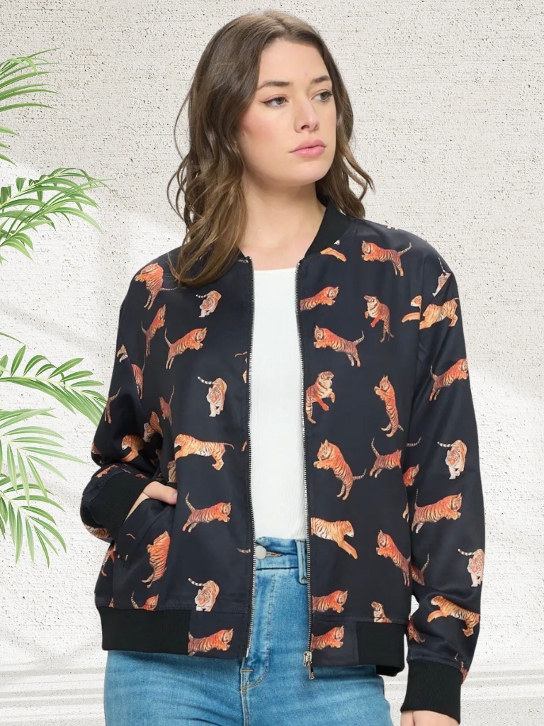A woman wearing a black bomber jacket with an all-over orange and brown tiger print, styled with a white top and blue jeans against a textured light gray wall with a green plant leaf on the left.