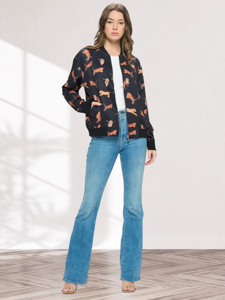A woman modeling a black bomber jacket with an orange and white dog print, paired with light blue flare jeans and a white top, standing in a brightly lit studio with a wood-patterned floor.