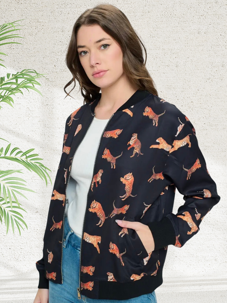 A woman wearing a black bomber jacket with an all-over orange and brown tiger print, styled over a white top and blue jeans against a textured light gray wall with a green plant leaf on the left.