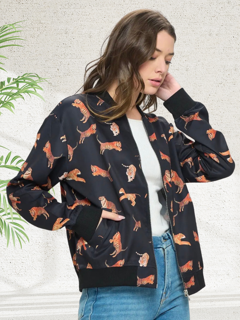A woman wearing a black bomber jacket with an orange and brown tiger print, styled with a white top and blue jeans against a textured light gray wall.